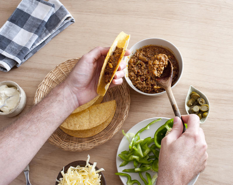 First person perspective view of hands filling a taco shell with ground meat beside green and hot peppers, lettuce and sour cream