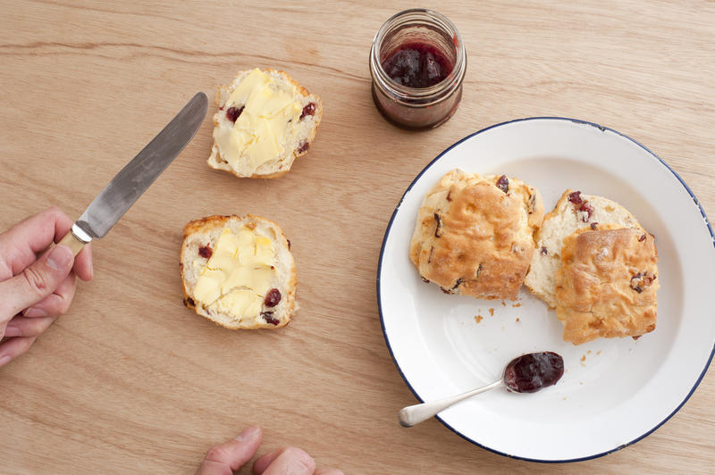 Man preparing fresh scones and jam for tea with a metal plate with two unbuttered scones alongside a halved scone with butter and jar of strawberry jam, overhead view with his hands and knife in view