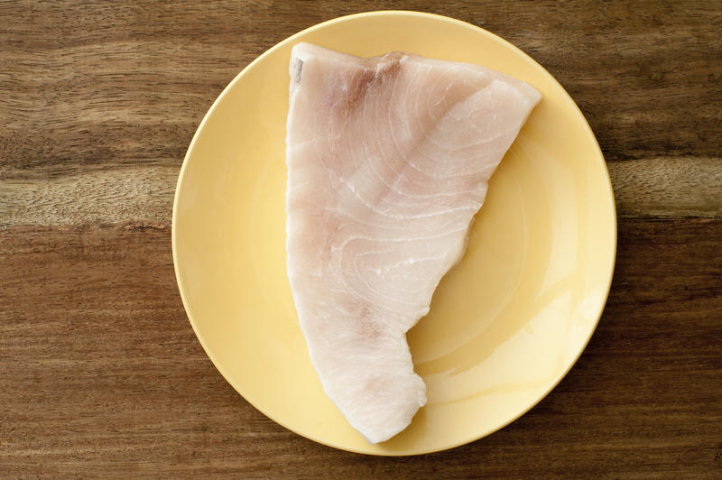 Top down view on raw portion of swordfish steak in simple round plate on brown wooden table surface