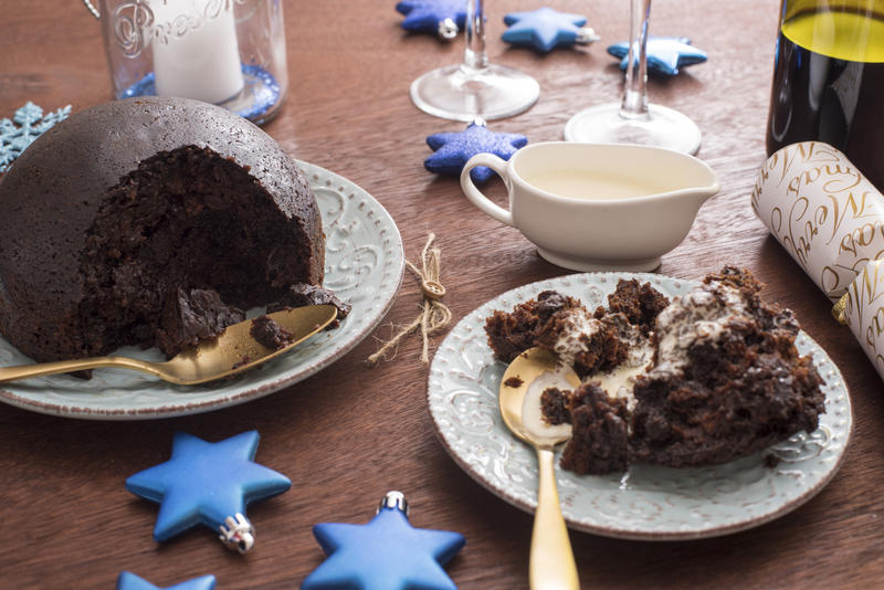 Serving of homemade fruity Christmas pudding topped with brandy cream on a festive dinner table with blue themed decorations