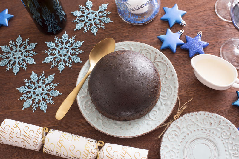 Delicious traditional fruity Christmas pudding waiting to be served on a festive table decorated with a cracker, wine, blue snowflakes and stars, overhead view