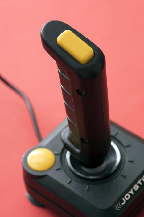 Classic stick gaming joystick of black plastic with yellow control buttons, close-up from above view against plain pink background
