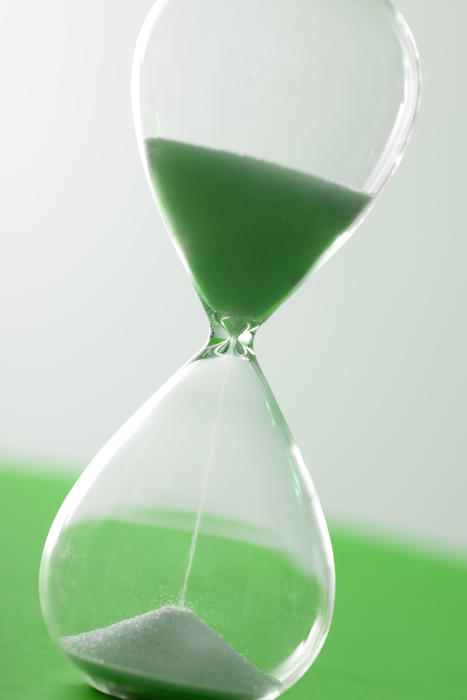 Single glass hourglass timer with pouring sand over green and white background for concept about time
