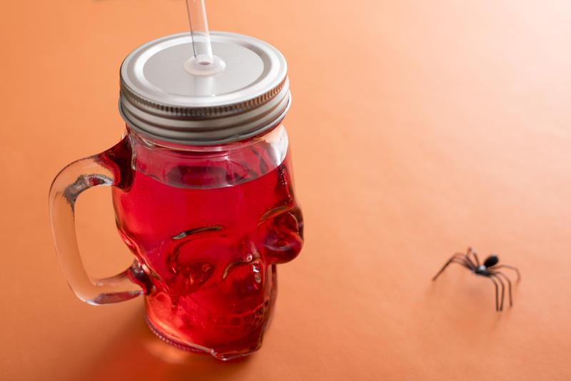 Red drink in glass shaped skull with large black spider posed beside it over orange background with copy space