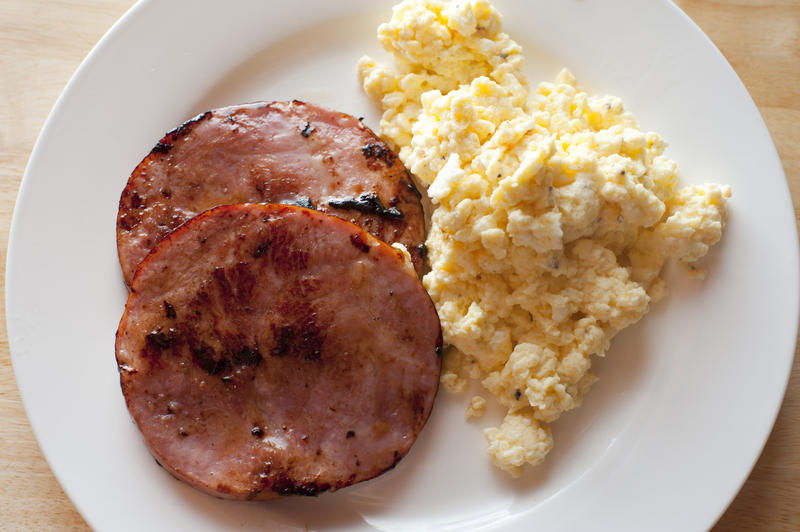 Two round shaped fried ham bacon slices beside hot scrambled eggs in white ceramic plate from first person perspective