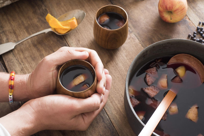 Person warming their hands on a mug of spicy fruity mulled wine seated at a table with a pot of freshly made brew