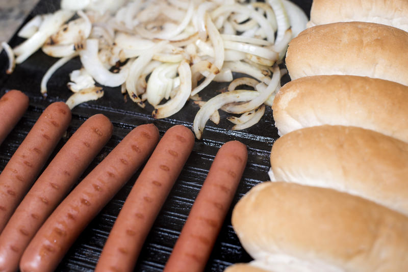 Several delicious hot dogs cooking on dark grill beside pile of sliced onions and buns