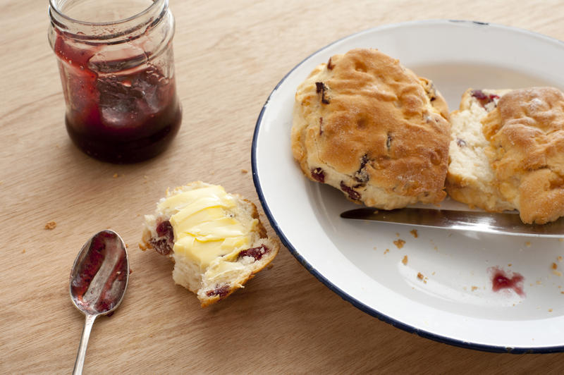 Table setting of partially eaten buttered crispy scone biscuits, messy spoon, plate and jam in plate over light brown wooden table