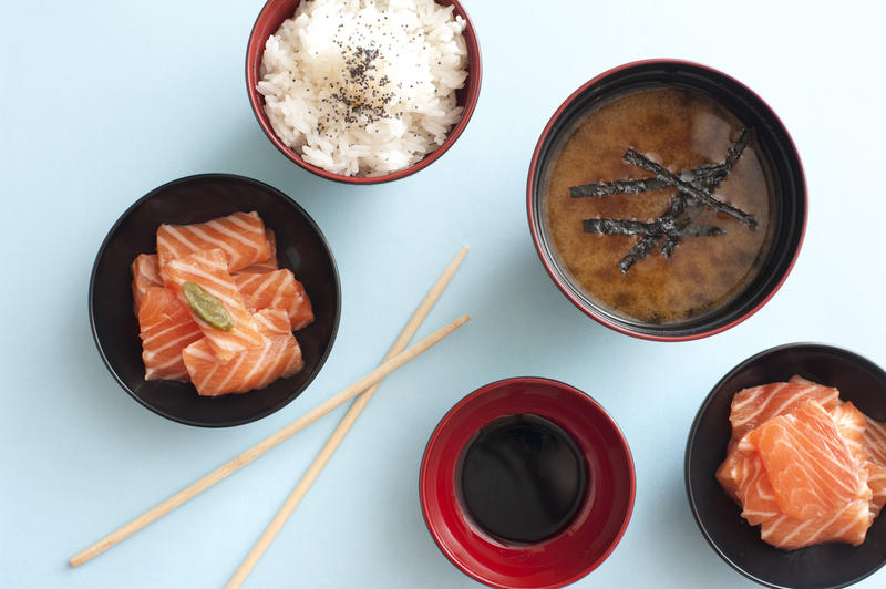 Overhead view of fresh cut and sliced salmon in small bowls beside soy dipping sauce and white rice by soup next to chopsticks against a light blue background