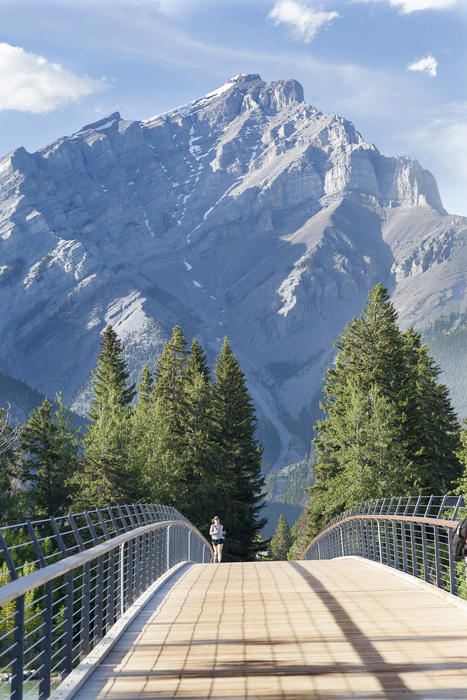 <p>A female jogger runs over a wooden bridge in Banff, Canada with the Rocky Mountains in the background</p>