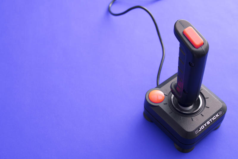 Classic joystick game controller of black plastic with red buttons and wire from above view on plain purple background with copy space