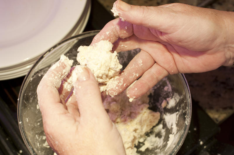 Hands shaping course dough into dumplings over glass bowl beside stack of white plates