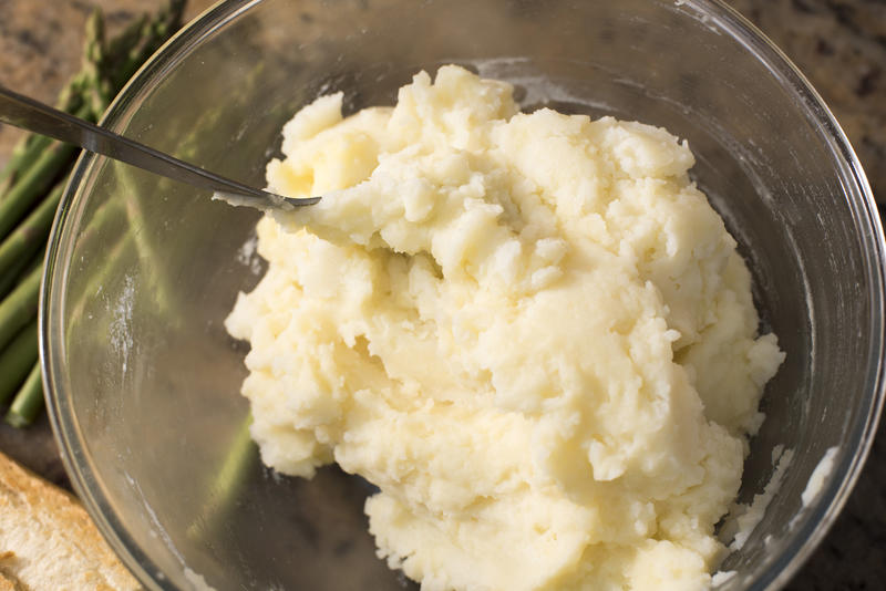 Buttered mashed potatoes in glass bowl with spoon set on a granite counter beside asparagus bunch