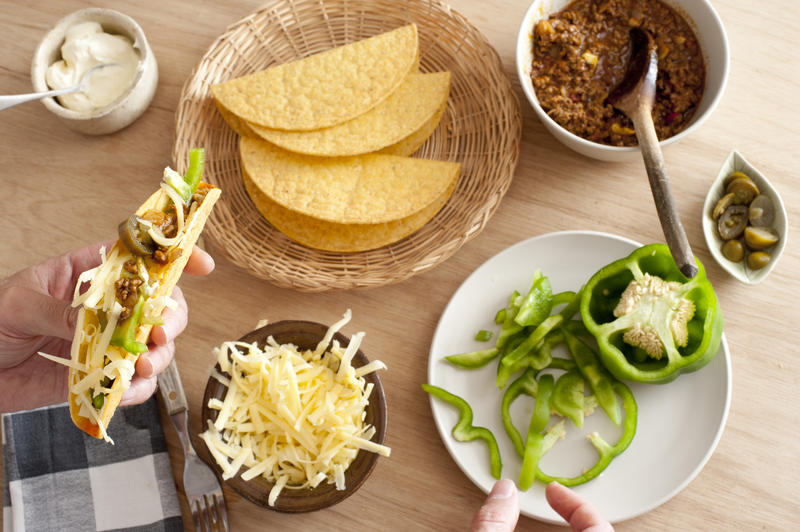 Overhead view of wood table set with ingredients to make hard shelled tacos including chopped green pepper, shredded cheese, and ground meat