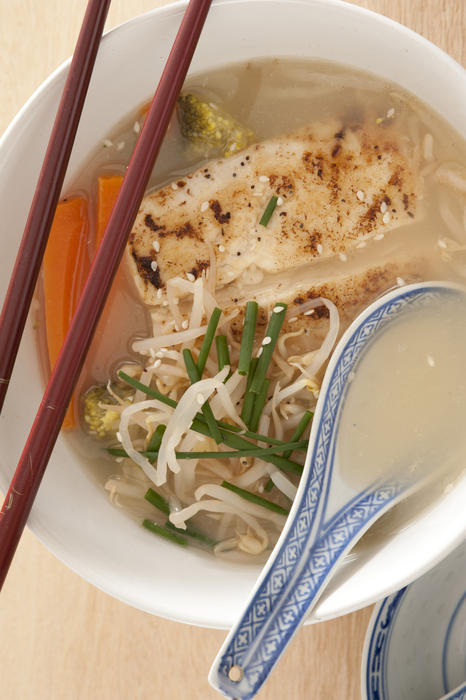 Top down view on milkfish noodle soup garnished with green chives, carrots and broccoli with brown chopsticks and ceramic spoon