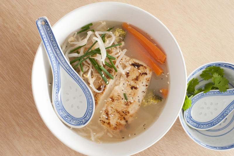 From above view on bowl of vegetables and fish meat and broth soup with spoon and garnishments with wooden surface background