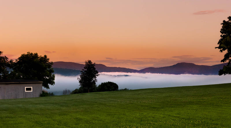 <p>Morning fog with shed and deep valley clouds contrasting sky and green grass with trees.</p>