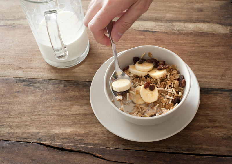 Single hand picking up spoon full of banana slice, raisin and muesli cereal in bowl beside half empty glass of milk over wooden table