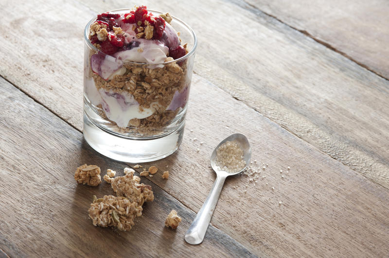 Glass filled with granola yogurt and fruit beside small silver spoon on rustic table