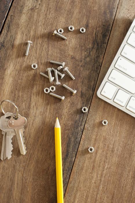 Nut and bolts lying on a wooden desk alongside a computer keyboard, pencil and keys in a conceptual image, overhead view