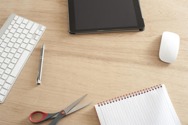 High Angle View of Desk with Scattered Office Supplies and Copy Space - Computer Keyboard and Mouse, Tablet, Scissors, Pen and Notebook on Wooden Desk with Central Blank Space