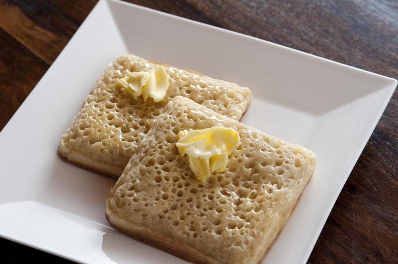 Two plain fried crumpets with butter on top neatly stacked partially over each other in simple matching square white plate