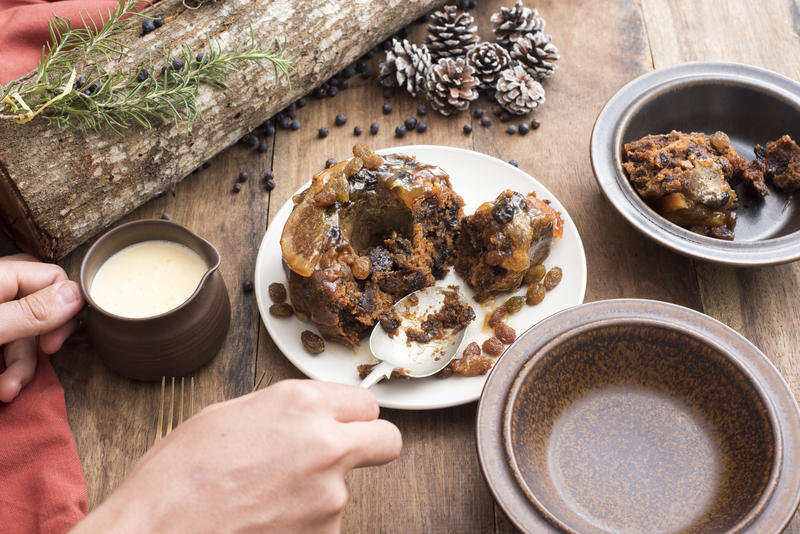 Man serving Christmas pudding and brandy sauce spooning it into two bowls on a rustic decorated table