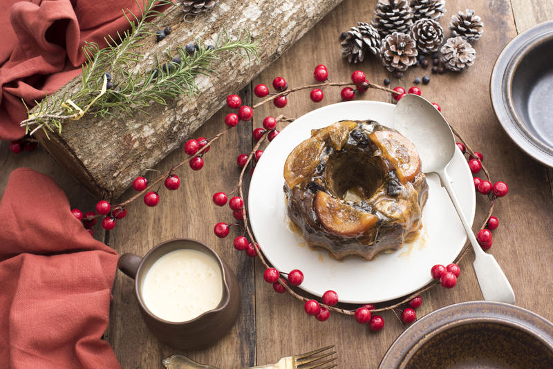 Tasty homemade fruity Christmas plum pudding with a jug of brandy sauce on a seasonal table with decorations viewed from above