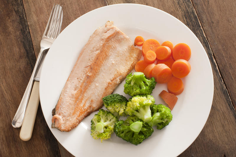 Single serving of baked rainbow trout next to steamed broccoli and sliced orange carrots in round plate over finished wood background