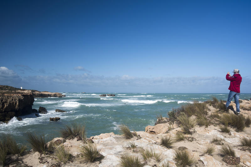 Man photographing the coastline at Robe, South Australia with rough seas breaking on rocky cliffs and a far distant view of the Obelisk