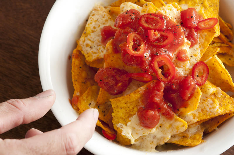 Man taking a nachos snack topped with melted cheese and hot chili and tomato salsa from a bowl on a buffet, close up view of his fingers and the food