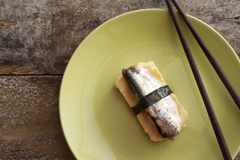 Single serving of sardine fish wrapped with strip of seaweed on plate with brown chopsticks over table