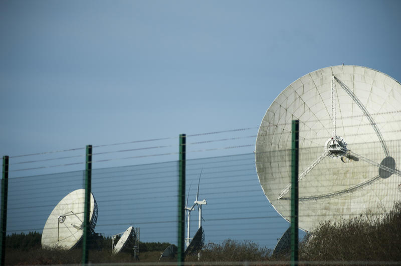 View of large satellite dishes or antennae for communications behind a high perimeter fence against a blue sky with copy space