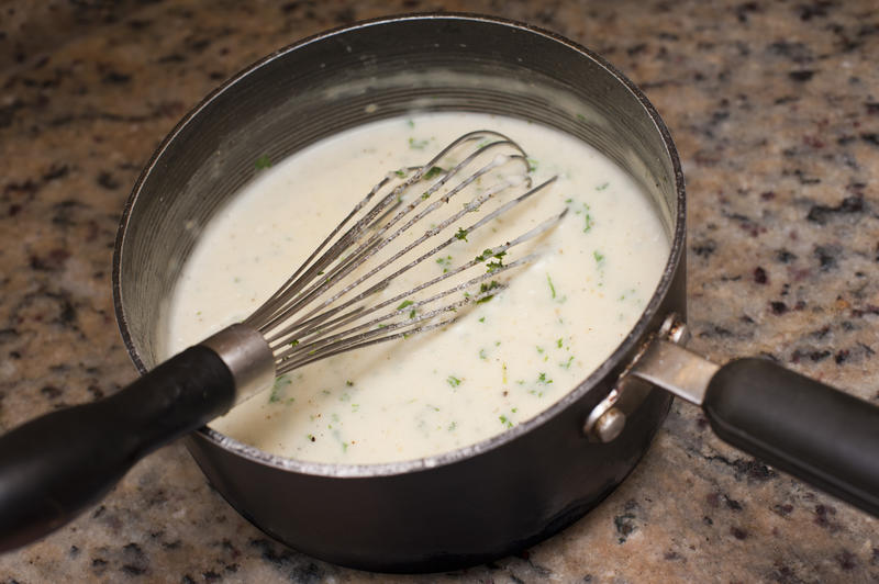 White creamy sauce seasoned with herbs in a saucepan with a metal hand whisk on a granite kitchen counter