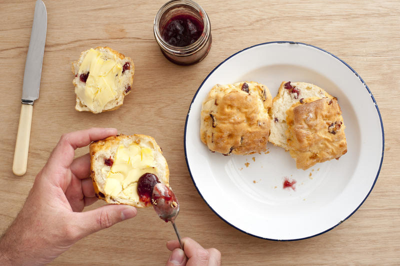 First person perspective view looking downward at hands coating two baked scones with butter, fruit jam and knife over wooden table