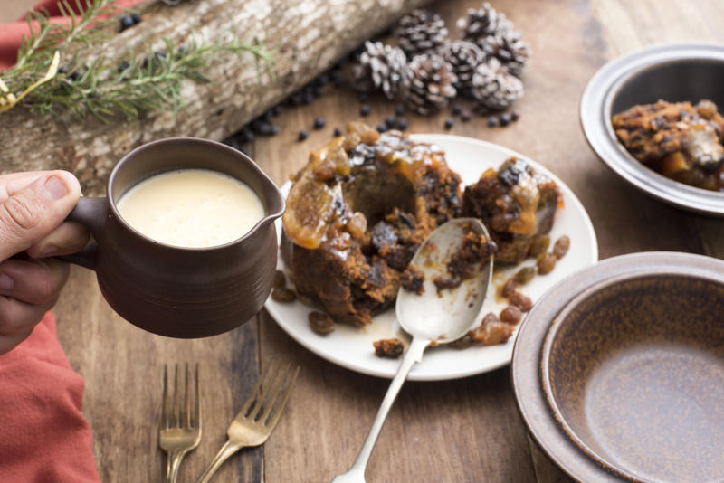 Person serving brandy sauce in a jug with traditional fruity Christmas plum pudding