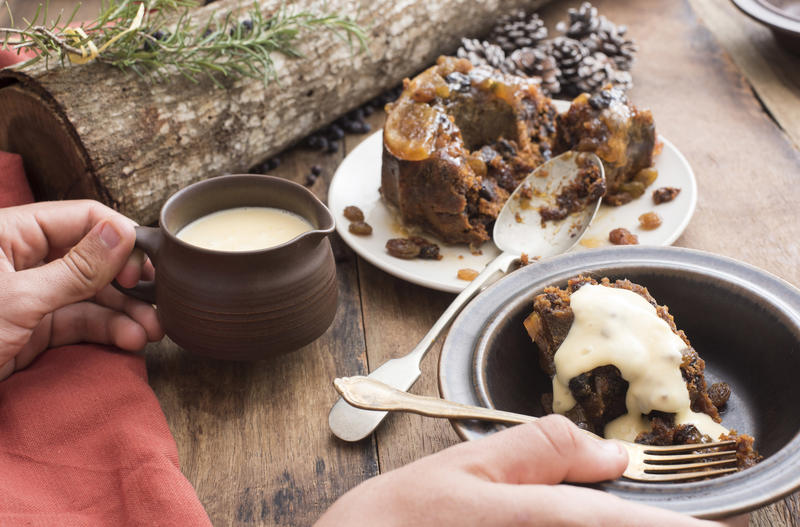 Person serving a helping of delicious fruity Christmas pudding topped with brandy sauce in a bowl at a decorated table