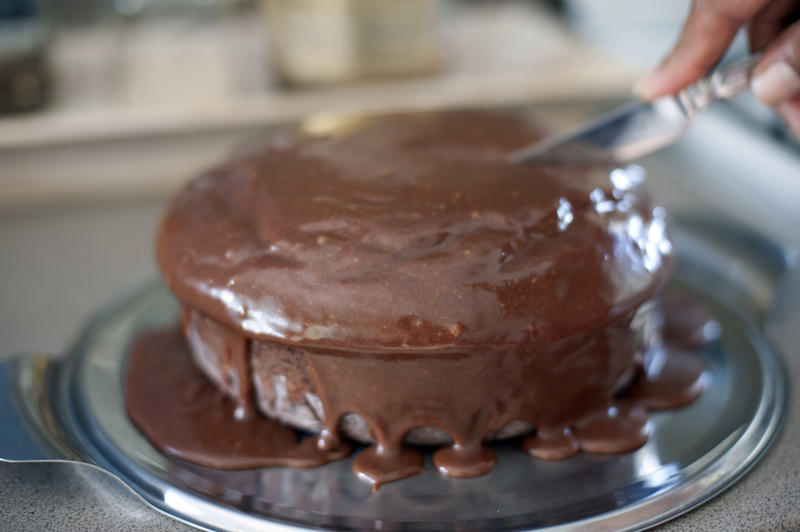 Out of focus knife cutting into chocolate cake with dripping icing over the sides on stainless steel tray