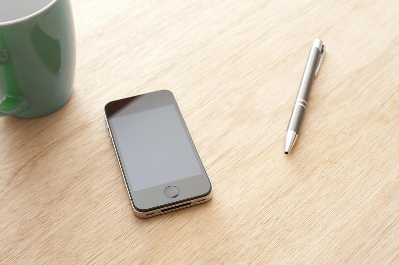 Touchscreen smartphone with black front panel on wooden table surface with pen and mug