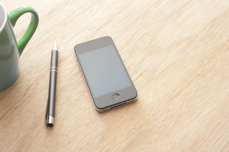 Smartphone with black pen and green mug on light wooden table surface, business communication concept with copy space