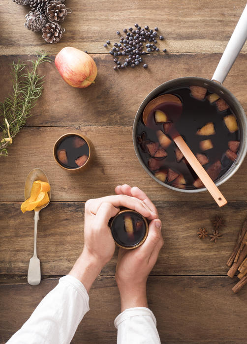 Man warming his hands on spicy hot mulled red wine for Christmas made with fresh fruit surrounded by the ingredients in an overhead view on a wood table
