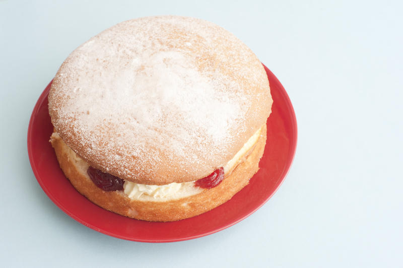 First person perspective top down view on single small size victoria sponge cake dessert in round red plate over light blue table with copy space