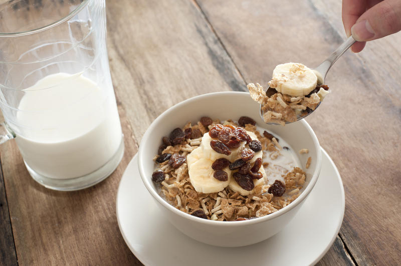 First person perspective view of spoon in hand reaching up to eat muesli granola cereal with slices of banana and raisins on top beside half empty glass pitcher of milk over table