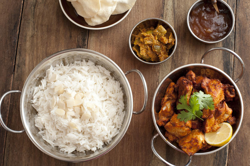 Overhead view of steamed rice and tandoori chicken besides chutney and fried bread set in stainless steel pots