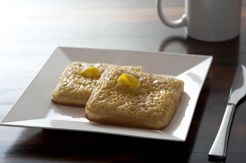 Two buttered crumpets in square plate beside stainless steel knife and coffee mug on dark wood table