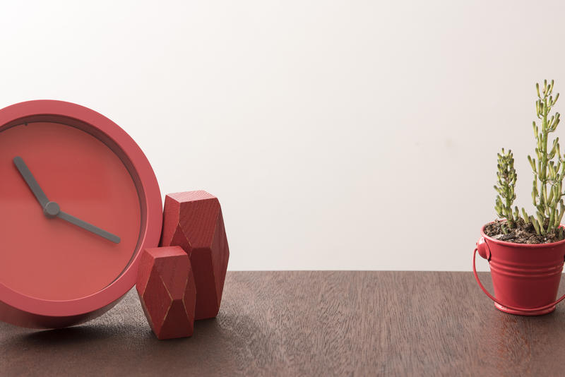 Red faceless clock on beside two polygonal shaped blocks on desk with little plant and copy space