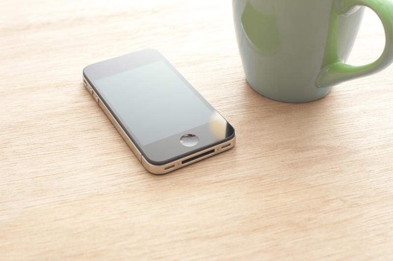 Touchscreen smartphone next to green mug close-up on wooden table surface. Copy space