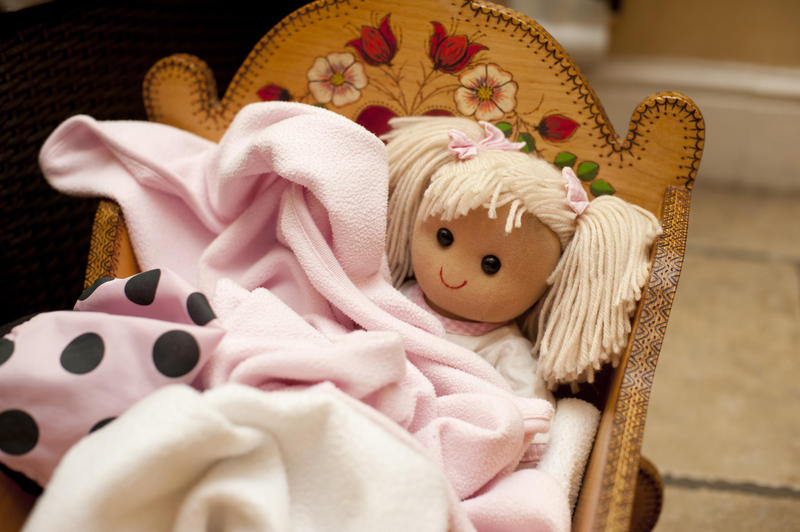 Cute little blond doll in a crib wrapped in a soft pink blanket in a kids playroom , close up high angle view