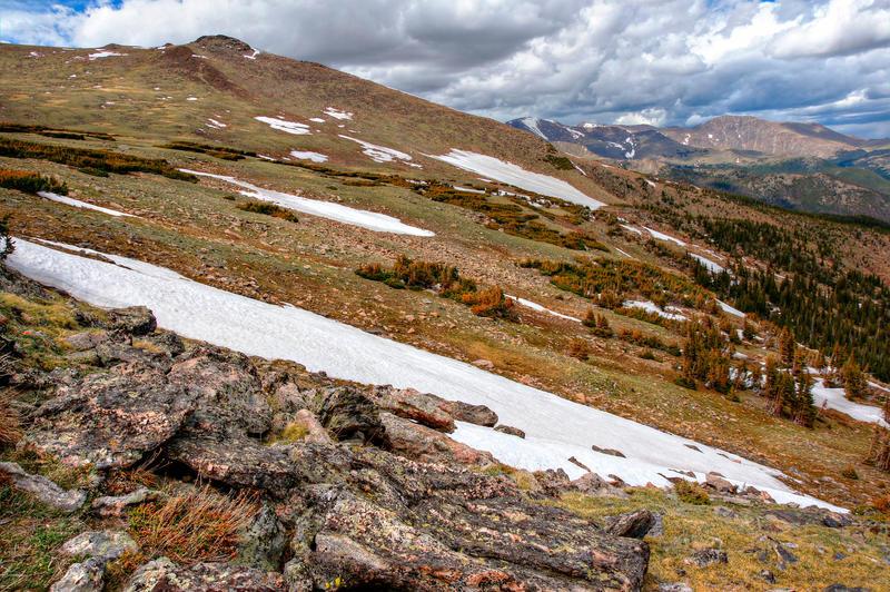 <p>Snow fields lie on the tundra at Rocky Mountain National Park. In this scene, rock in the foreground gives way to a series of high altitude pine forests and snow fields. Distant mountains lie under stormy clouds.</p>