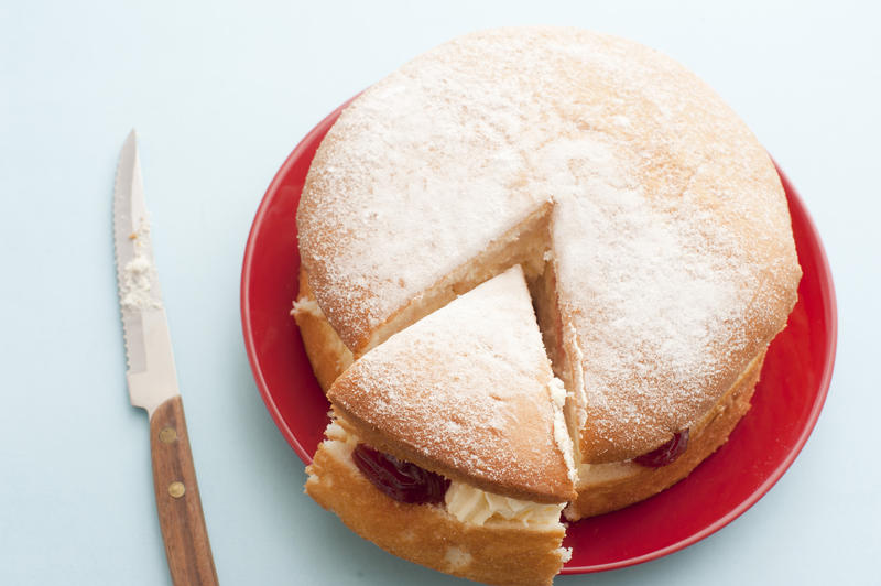 Delicious fresh sponge cream cake with strawberry jam sliced for tea on a red plate, high angle view with knife alongside